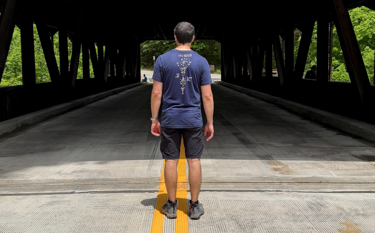 A man stands with his back to the camera in the center of a covered bridge roadway, wearing a navy blue t-shirt with a white graphic print on the back, dark shorts, and gray sneakers.