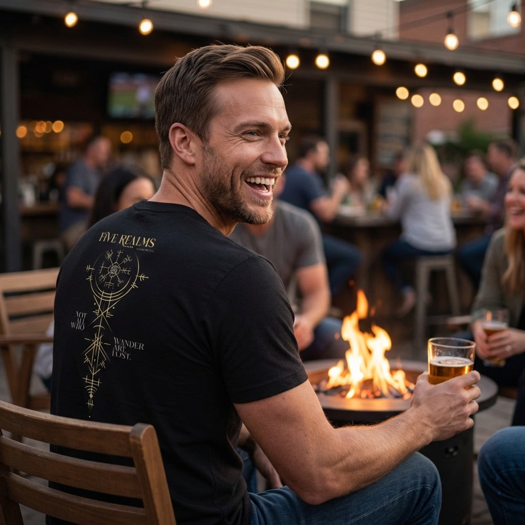 Man sitting outdoors at a fire pit, holding a beer, with a casual atmosphere. He wears a black T-Shirt with a printed nordic compass design on it.