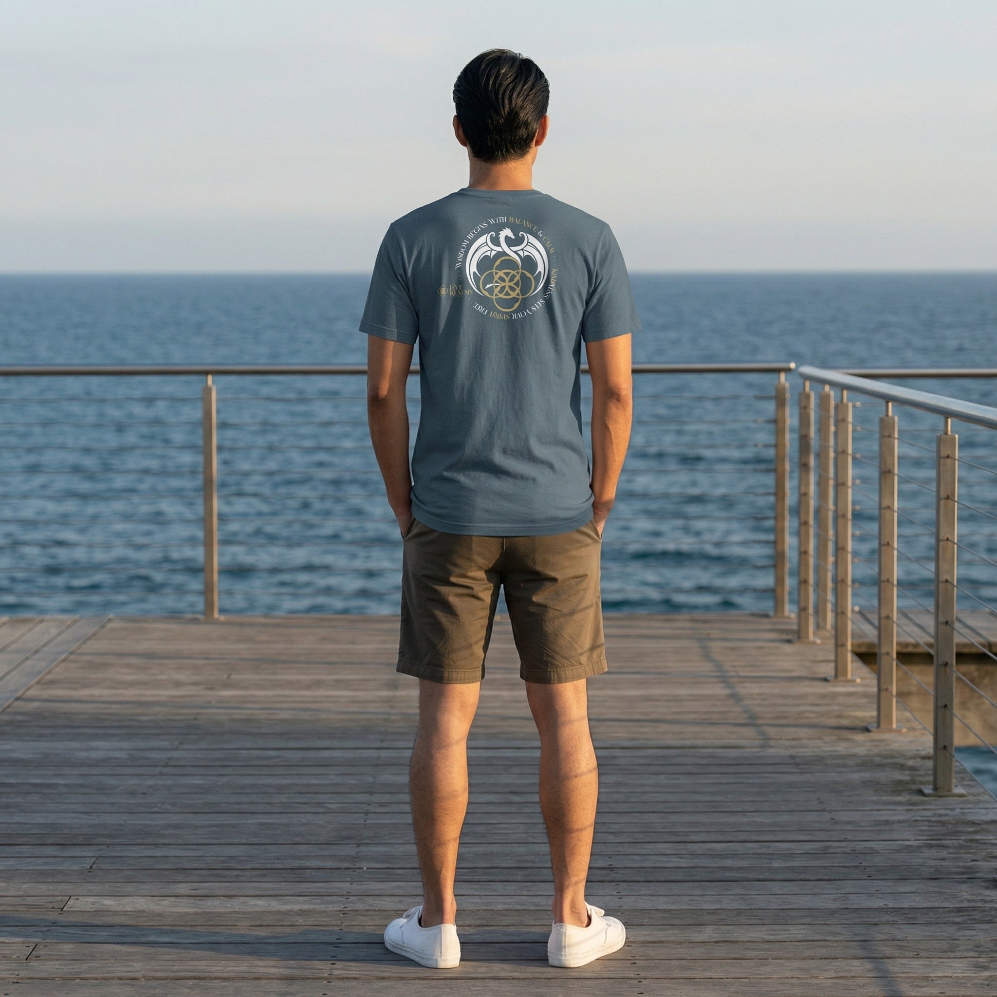 Man standing on a wooden pier overlooking the ocean, wearing a gray t-shirt with a logo.