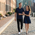 Man and woman walking along a waterfront street with buildings and water in the background. Man wears a dark blue polo and black pants. Woman wears a dark blue dress.