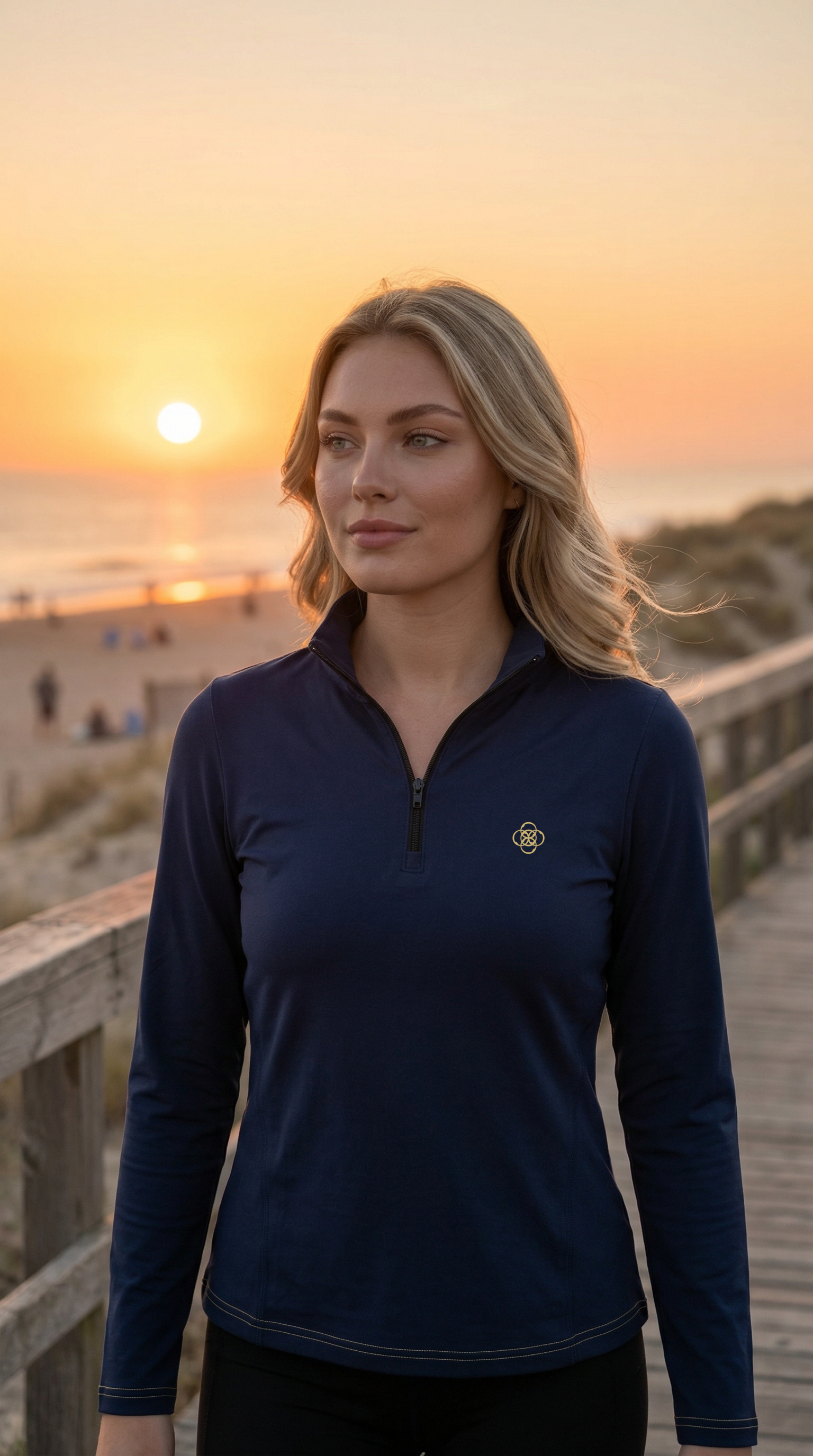 Woman wearing a navy blue pullover with a logo on a wooden boardwalk during sunset.