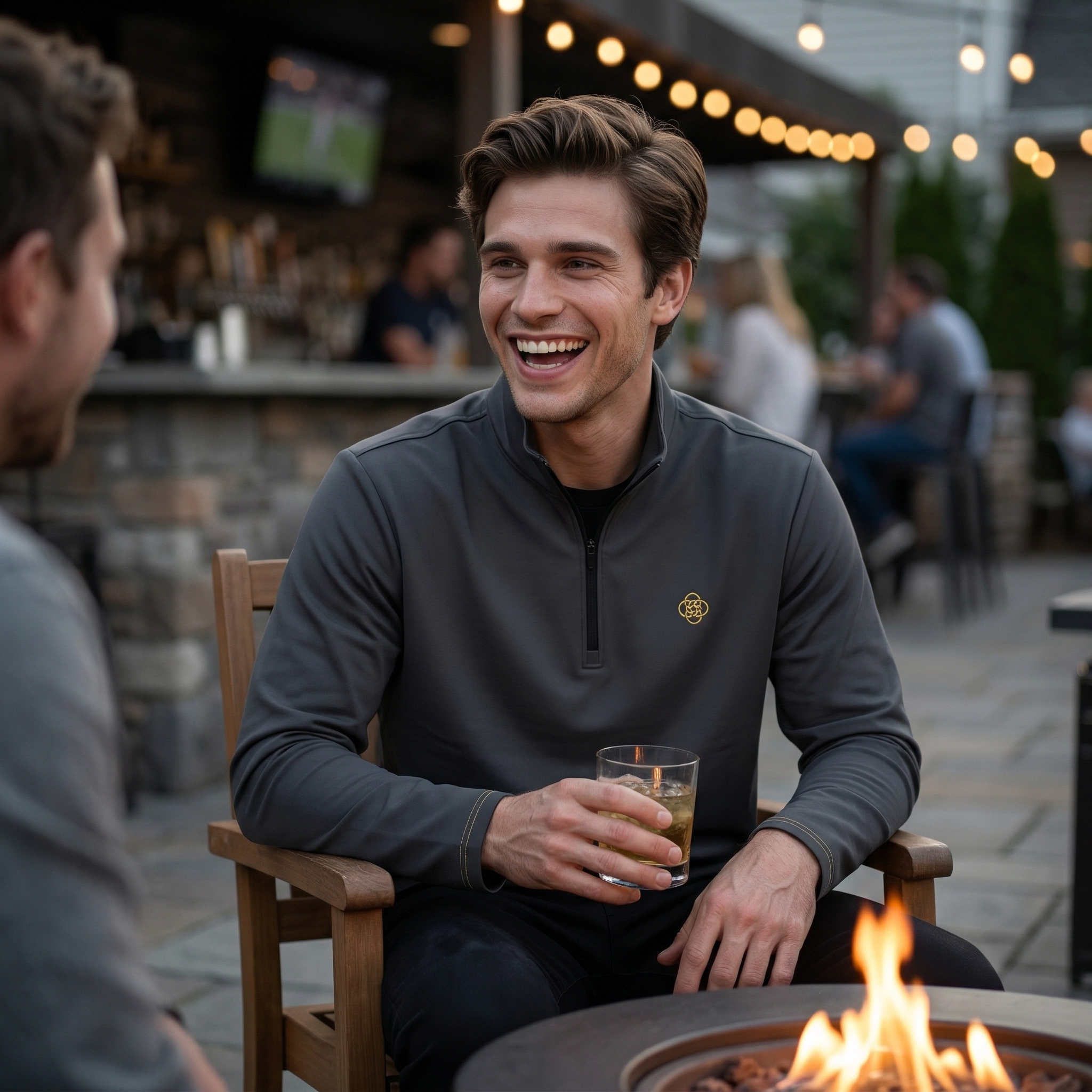 Man sitting by a fire pit holding a drink, smiling at another person outdoors. He wears a dark grey quarter zip with the Five Realms logo on it.